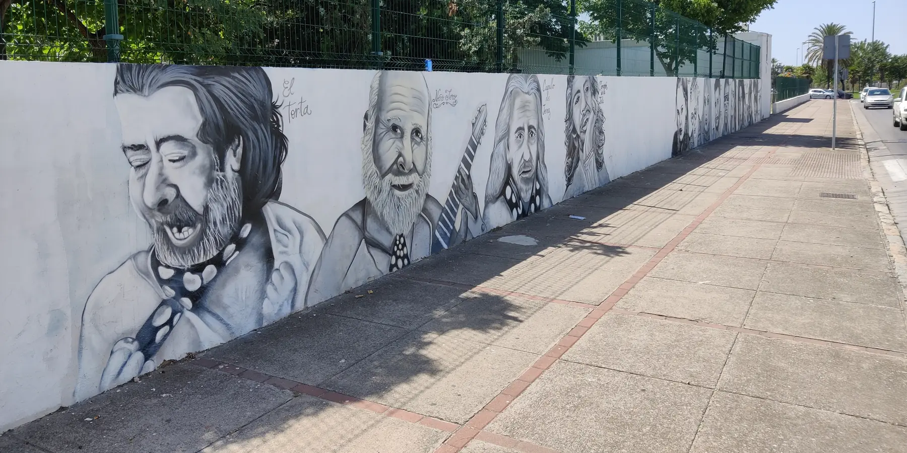 A long whitewashed school wall in Jerez painted with grey portraits of flamenco singers and a guitarist, seen along a pavement under trees
