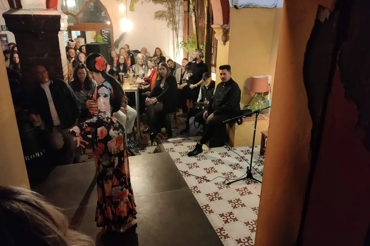 A dancer in a flowered dress turns on a low stage beside a seated guitarist while an audience sits packed in a tiled courtyard