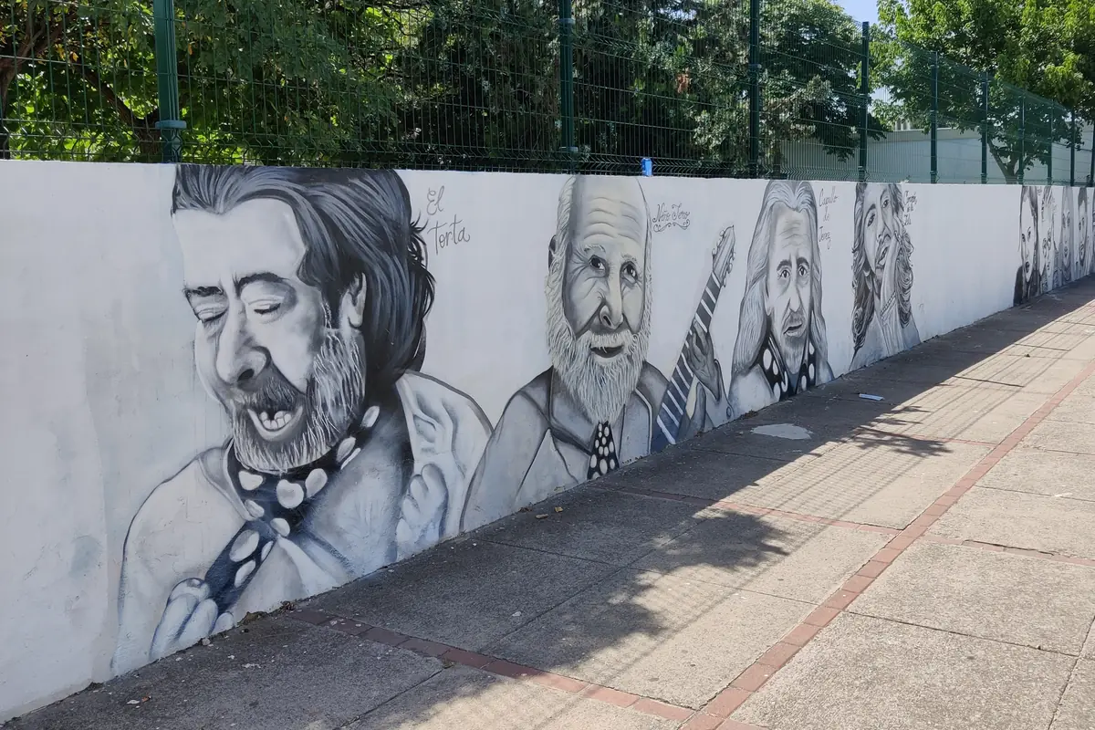 A long whitewashed school wall in Jerez painted with grey portraits of flamenco singers and guitarists, beside a pavement