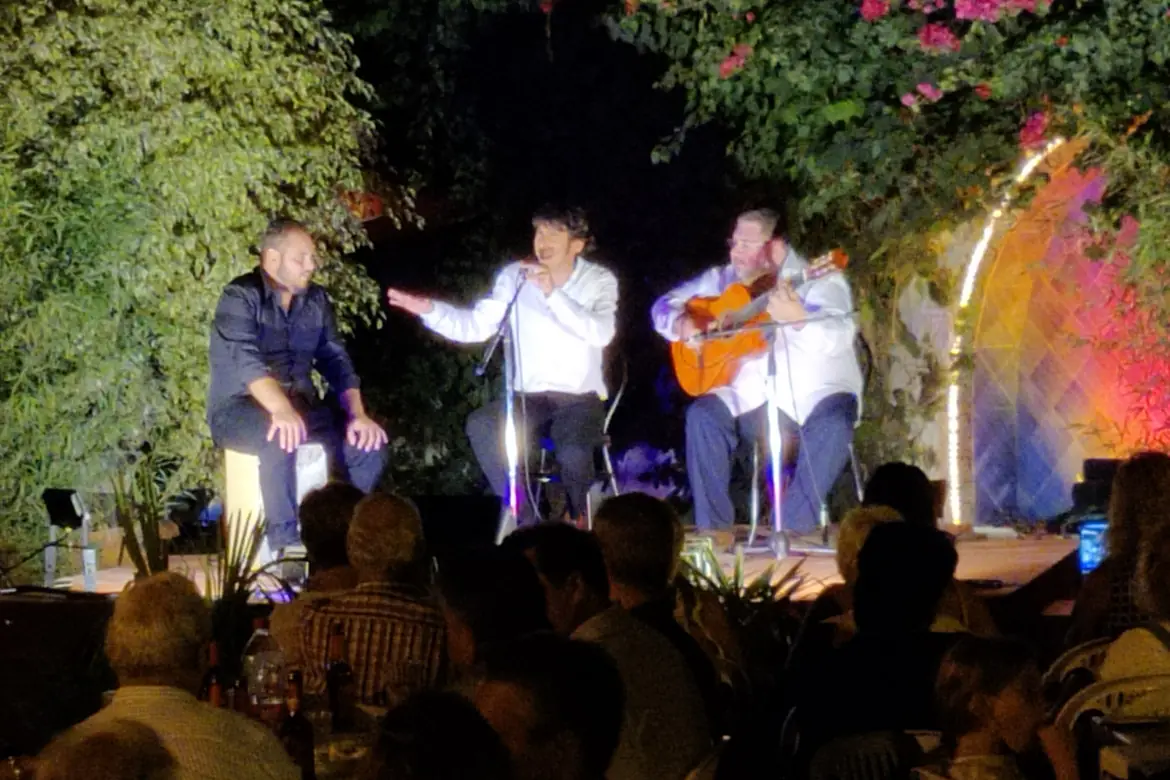 A singer in a white shirt gestures between a seated musician and a guitarist on a night stage with flowering vines behind them